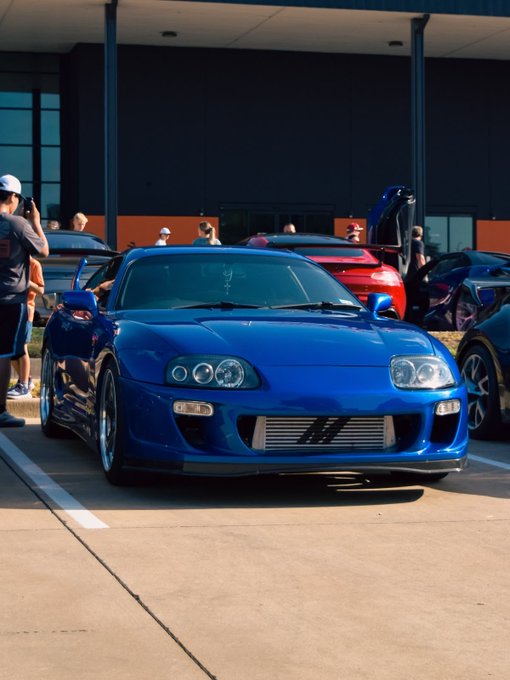 A bright blue modified sports car (appears to be a Toyota Supra) parked at what looks like a car meet, with people in the background and other vehicles visible.
