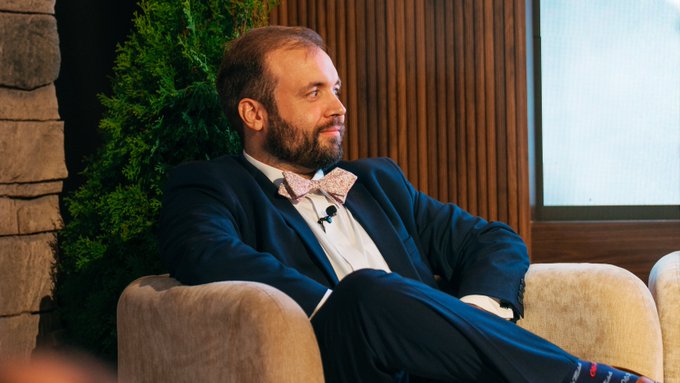 A bearded man in a navy suit with a pink patterned bow tie sits relaxed in a tan armchair, smiling off to the side during what appears to be a panel discussion or interview. The post "Mentally, I'm here:" paired with the image humorously conveys a sense of being checked out or daydreaming despite appearing engaged in the professional setting.