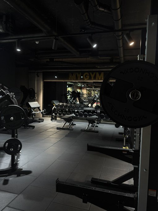 A dimly lit interior of a gym called 'MYGYM', featuring weightlifting equipment in the foreground including plates labeled 'MASONHOE TECHNOLOGY 20' on a barbell rack, with a wall of dumbbells visible in the background.