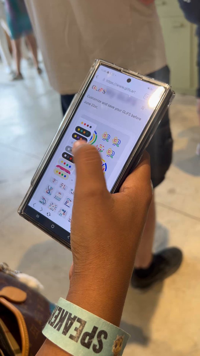 The image shows a person holding up their phone displaying a colorful app with a cartoon character on it. The person's hand is wearing a green wristband that says "SPAM" on it, indicating they might be participating in an event or competition related to spam emails. The background of the image features people walking by and chairs placed around them, suggesting this could be taking place at an event or gathering where participants are engaged with various activities.