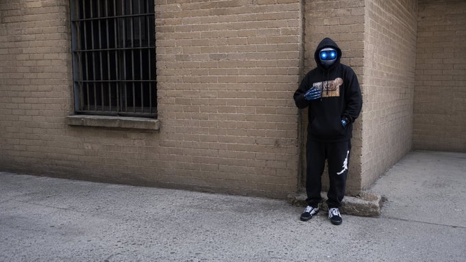 A person wearing a black hoodie with glowing blue goggles stands in an urban alleyway between brick walls, placing a hand on their chest.