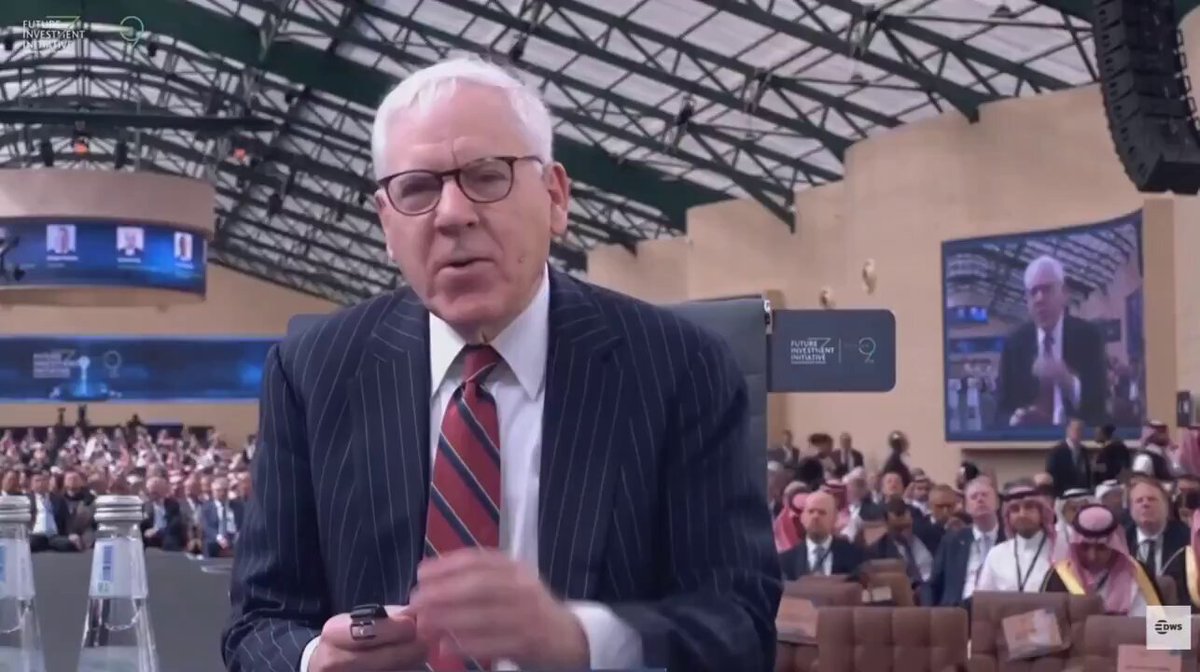 The image depicts an older man wearing glasses, dressed in a suit, standing next to a large screen displaying two news articles. He appears to be engaged in a discussion or presentation about the stock market, with the backdrop of an indoor event space featuring multiple people sitting and others standing nearby. The post behind him reads: "WALL STREET WARNS OF FIAT DEBASEMENT, BACKSBITCOIN $14 trillion BlackRock CEO says Bitcoin is a hedge against "fiat debasement". This suggests that the older man might be speaking at or near an event related to cryptocurrency and its potential impact on traditional financial systems. The presence of multiple people around him further indicates a communal gathering focused on discussing or presenting new ideas, especially in relation to emerging technologies like Bitcoin.