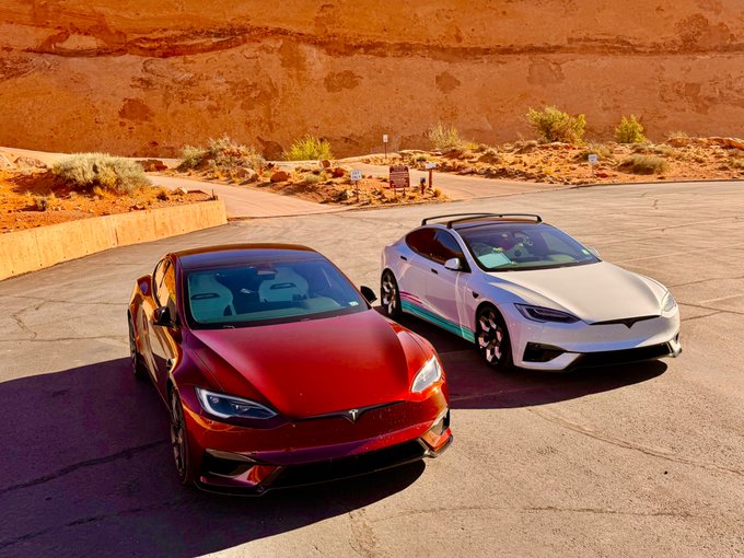 Two Tesla Model S Plaid cars, one red and one white, are parked side-by-side on a paved surface against a backdrop of red rock cliffs.