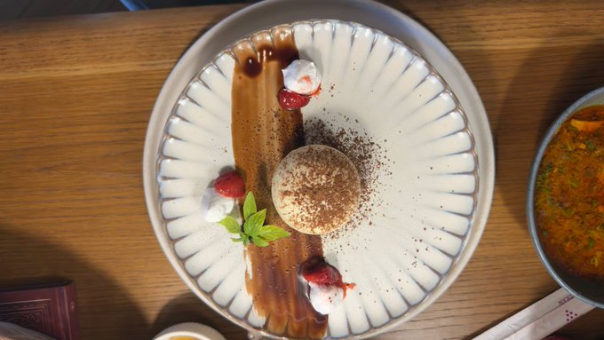 An overhead view of a beautifully plated dessert on a white scalloped plate, featuring a round chocolate-dusted cake, cream dollops with strawberries, mint leaves, and a caramel drizzle.
