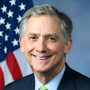 A portrait of a smiling man in a dark suit with a green tie stands before an American flag. The image likely depicts French Hill, the House Financial Services Chair mentioned in the post announcing a field hearing on the CLARITY Act in New York on July 17.