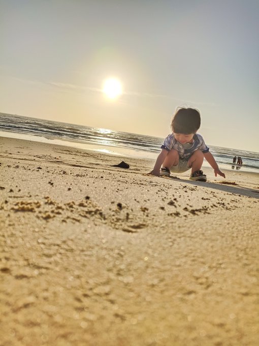 The image shows a young child playing at the beach with sand between their toes. The sun is setting behind them, creating a beautiful backdrop for this scene. The photo has been edited to focus solely on the child's activity of digging in the sand, capturing the essence of a fun and carefree moment spent by the ocean.