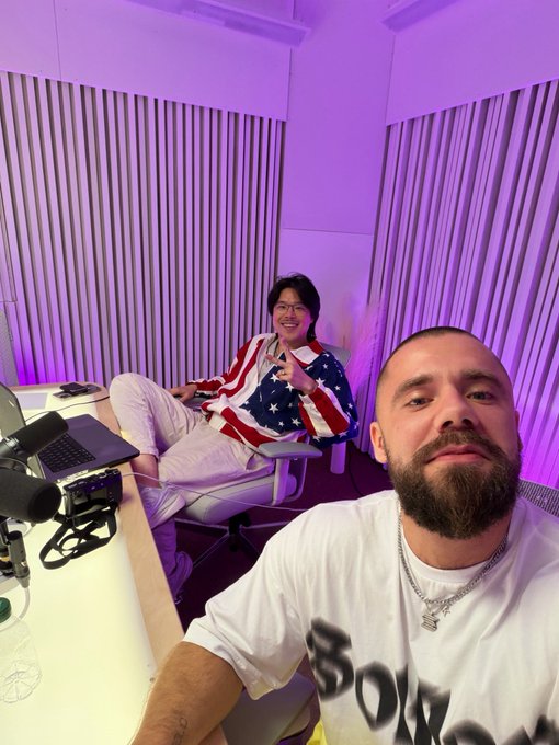 Two men in a room with purple lighting; one sits in a chair wearing an American flag-patterned shirt, while the other takes a selfie wearing a white t-shirt with black graffiti-style lettering.