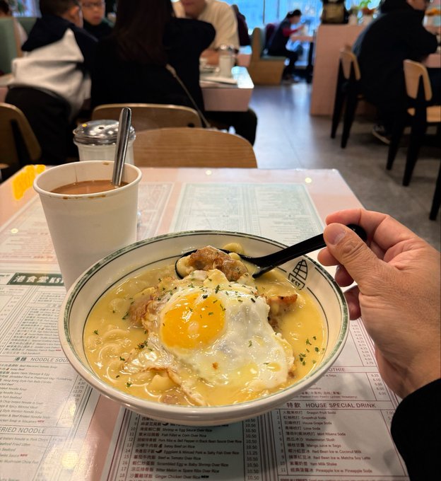 A person holds a spoon over a bowl of creamy soup topped with a sunny-side-up egg, accompanied by a side of coffee or tea at a noodle soup restaurant, with the menu visible underneath. The cozy meal reflects the "power up with a good breakfast" sentiment in the post, showcasing a hearty start to a Saturday before heading out for the day.