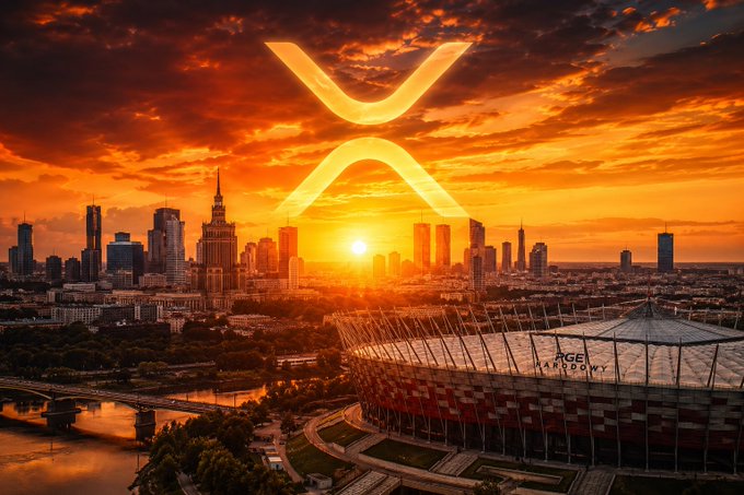 The image features a city skyline with an XRP logo at sunset over the city. The sun is setting behind the buildings, creating a warm glow that illuminates the scene. The sky is filled with orange and yellow hues as the sun sets in the background. In the foreground of the photo, there are two buildings prominently visible - one large stadium and another smaller building. The XRP logo stands out against this backdrop, drawing attention to its presence.