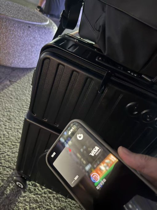 A hand holding a smartphone displaying a Visa card on the screen, positioned in front of a black hard-shell suitcase in what appears to be an airport or travel setting.