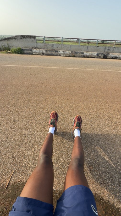 Person extending their legs forward on a road, wearing orange running shoes and white socks, with a bridge visible in the background, capturing a post-run stretch that aligns with the "jog maxxing" theme of maximizing running performance.