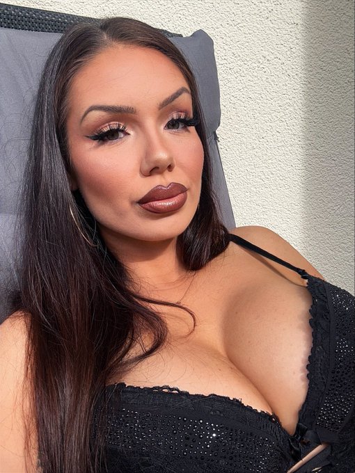 A woman with long dark hair poses for a close-up selfie wearing a black lace-trimmed top, displaying heavy makeup with bold brown lipstick and large hoop earrings.