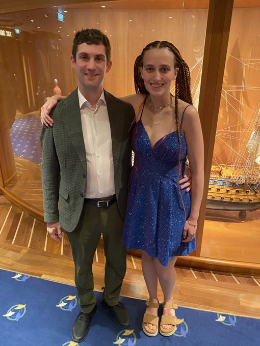 A photograph of a couple posing together, with the man wearing a green blazer and white shirt and the woman in a sparkly blue dress, standing in front of a glass display containing a detailed model ship.