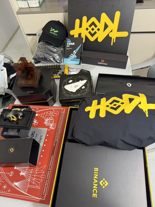 A collection of Binance and HODL merchandise and branded items including t-shirts, a cap, a plaque, and gift boxes spread on a red Christmas-themed mat.