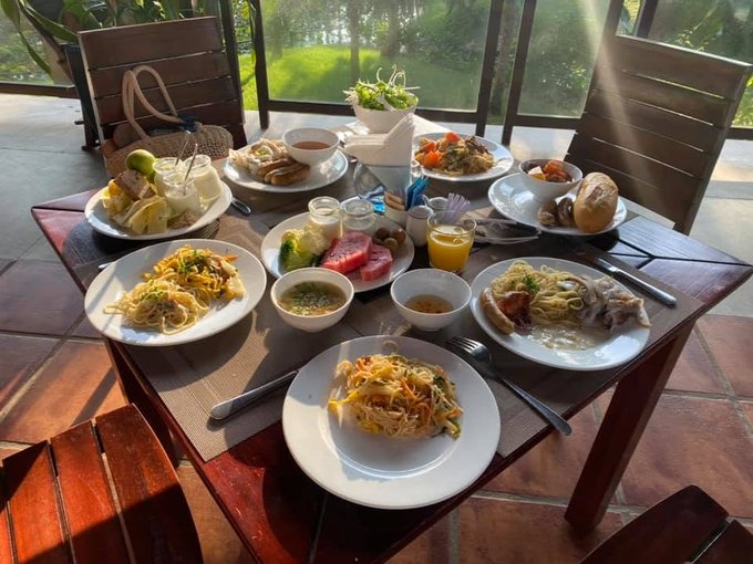 A lavish breakfast spread on a wooden table including multiple plates of pasta/noodles, sliced watermelon, fresh fruit, eggs, salad, juice, and bread, set in an open-air dining area.
