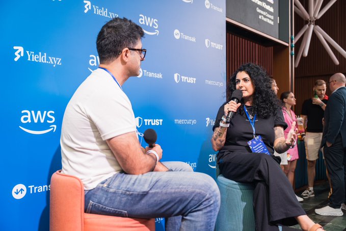 Two people are seated and engaged in a conversation at a conference, with one holding a microphone. The backdrop features logos for Yield.xyz, AWS, Transak, Trust, and Mercuryxo.