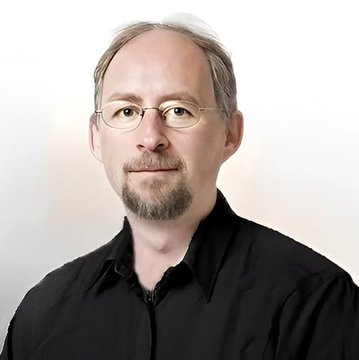 A professional headshot of a man with glasses, a goatee, and short gray hair, wearing a black collared shirt against a plain white background.