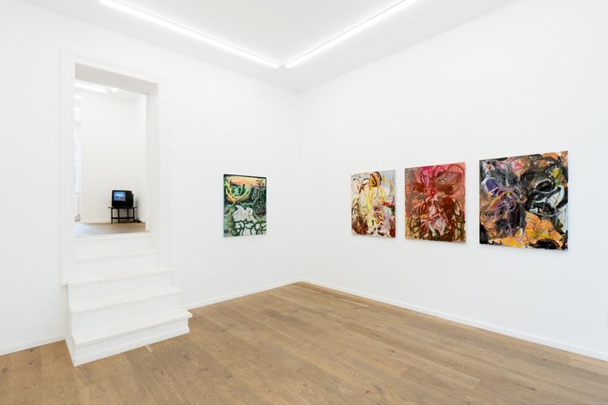 A bright white art gallery interior with light wood flooring, a staircase leading to an upper level, and four colorful abstract paintings displayed on the right wall.