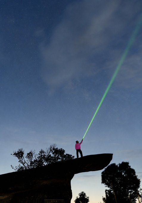 A silhouette of a person in a pink jacket standing on a rock formation at night, shining a bright green laser/light into the starry sky.