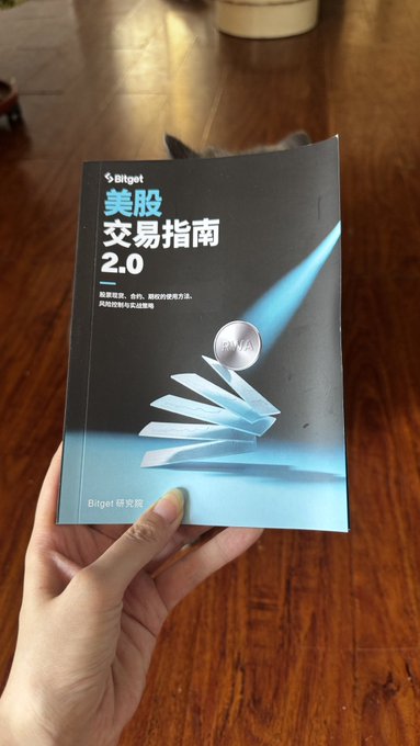A hand holds a Bitget '美股交易指南 2.0' (US Stock Trading Guide 2.0) brochure showcasing stock market navigation graphics.
