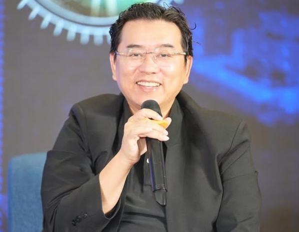 A photograph of a smiling man in a black suit and glasses speaking into a microphone against a blue backdrop with gear imagery.