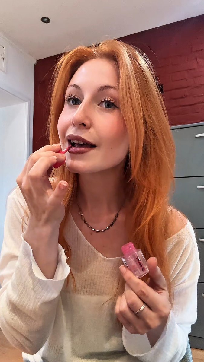 A young woman with reddish-blonde hair is applying lip gloss in front of a mirror, wearing a white ribbed top and a gold chain necklace.
