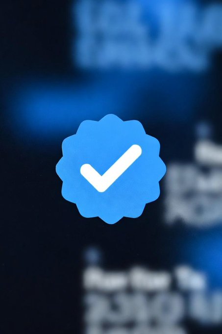 A close-up of a blue scalloped verification badge with a white checkmark, displayed on what appears to be a Twitter/X profile screen.