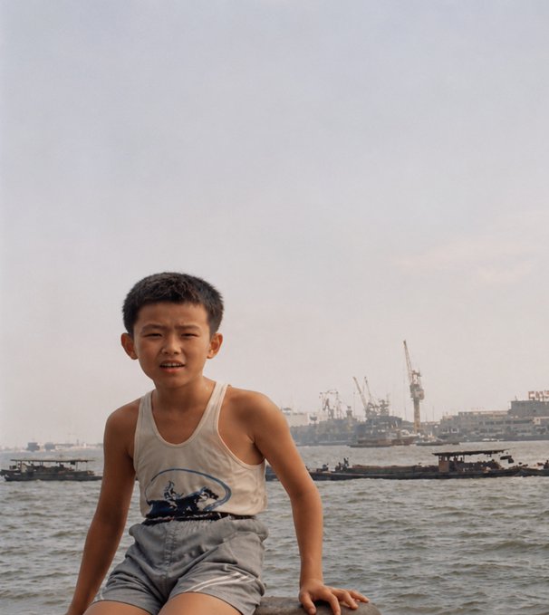 A young boy with a scrunched expression sits by a harbor with cranes visible in the background. The image appears to be an old photograph, humorously captioned as a childhood photo of CZ (Changpeng Zhao), the Binance founder and billionaire, suggesting this is him as a child.