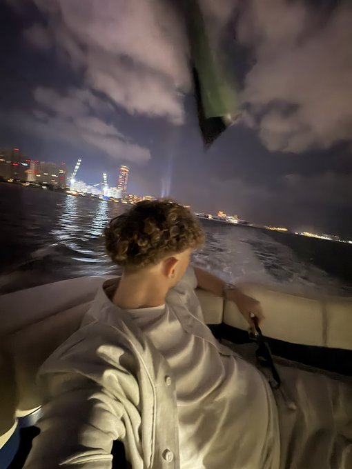 A man with curly blonde hair sits on a boat at night, wearing a cream-colored hoodie while looking out at a city skyline illuminated across the water. The moody, atmospheric scene with clouds and city lights pairs with "GN" (good night), capturing a serene moment before bed.