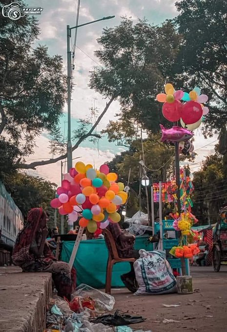 A street scene showing a vendor selling colorful balloons and party supplies, with a person in a red headscarf sitting on a low wall to the left near a train track, with trees and power lines in the background.
