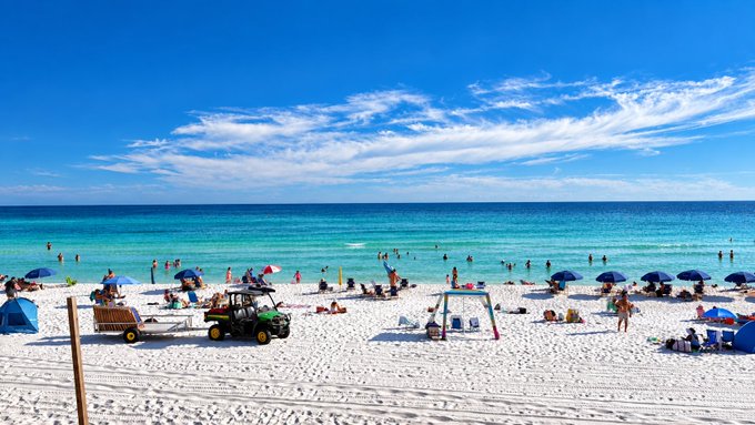A vibrant tropical beach scene with crystal-clear turquoise water, white sand, numerous beachgoers with colorful umbrellas, a green utility vehicle on the sand, and bright blue skies with scattered clouds.