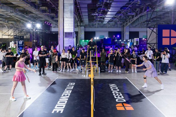 Two female players face off in a badminton match at a badminton event, with the court labeled 'DEERMODE' and 'DEEPCON' while a crowd of spectators watches from behind.