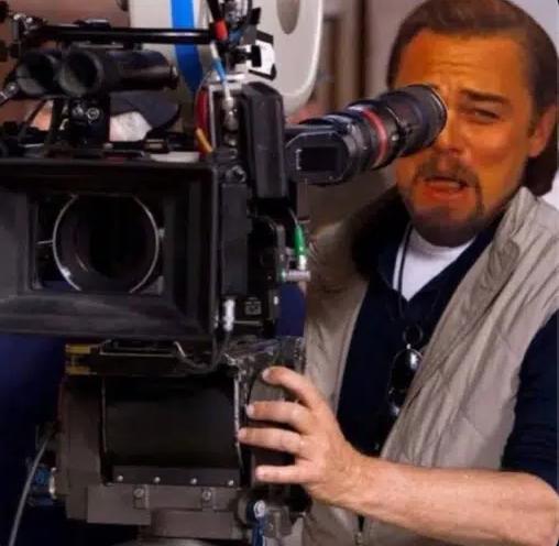 The image features a man wearing a vest, holding a camera up to his face with one hand while using the other hand to adjust the lens. The man appears to be focused on something outside of the frame, possibly a subject or an object that has caught his attention. The caption "Perez looking at this guy like:", which is likely a reference to the actor who played the character "Pepsi" in the movie "The Social Network".