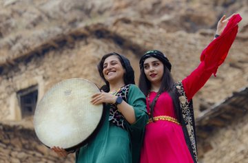 Two women in vibrant traditional clothing—one in green and the other in red with a decorated headdress—posing cheerfully with a large drum against a stone wall background.