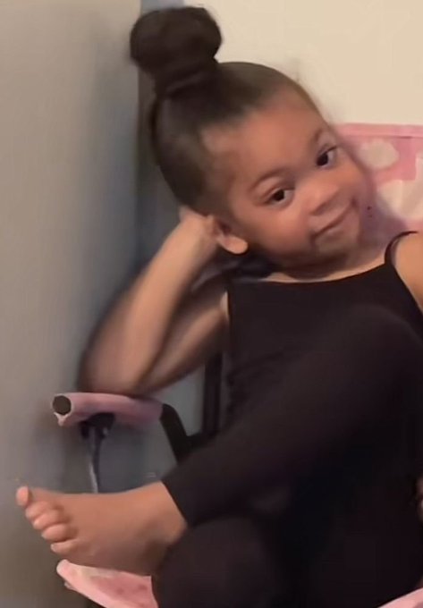 A young girl with her hair in a high bun, wearing a black tank top and black pants, sitting against a wall and smiling at the camera.