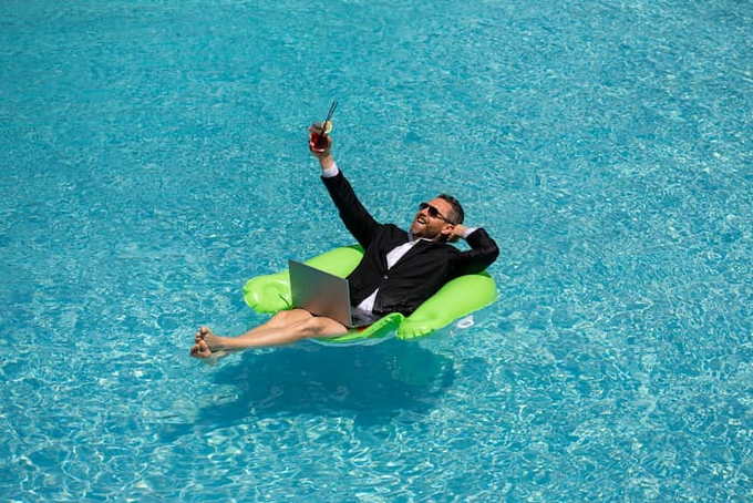 A businessman in a tuxedo and sunglasses lounges on a green inflatable float in a turquoise pool, holding up a drink in a celebratory toast while working on a laptop, perfectly capturing the post's message of "No one Intern finally gets an Intern: Agent OS."—a humorous depiction of effortless, autonomous assistance that lets him relax while his AI "intern" handles the work.