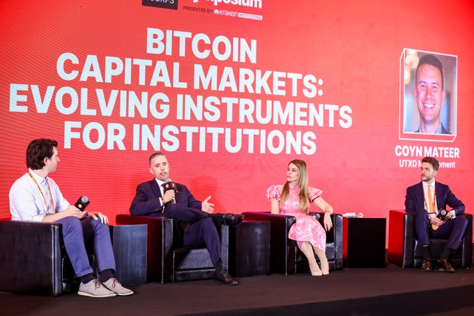 A panel discussion on stage with the title 'BITCOIN CAPITAL MARKETS: EVOLVING INSTRUMENTS FOR INSTITUTIONS' on a large red screen, featuring four seated participants and a photo of 'COYN MATEER UTXO Management'.