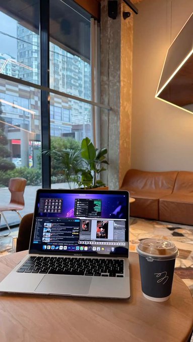 A laptop displaying a coding interface and a paper coffee cup rest on a wooden table in a modern cafe, with a brown leather sofa and large windows in the background.
