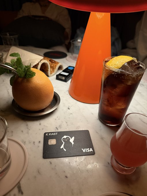 A marble table setting featuring a black 'KAST' Visa credit card with a ghost design, alongside a citrus drink, an orange cocktail in a hollowed orange, and a tall orange lamp.