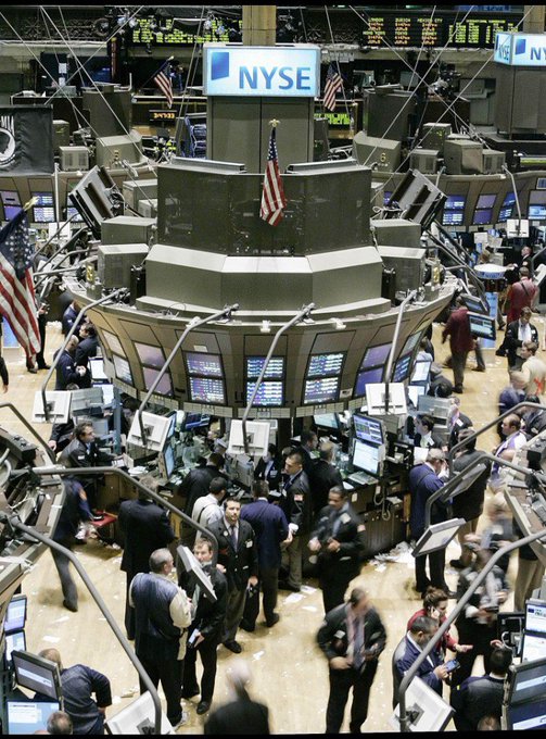 A high-angle view of the New York Stock Exchange (NYSE) trading floor, showing the central podium with an American flag and the NYSE logo above, surrounded by traders, monitors, and stock data boards.