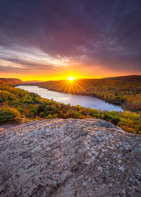 The image shows a beautiful sunset over a lake surrounded by trees with orange leaves. The sun is setting behind the mountains, creating a stunning backdrop for the scene. The photo has been captioned "GM GQuack Fans. Have a perfect day. Take a sip of your coffee. Keep the conversation going about @wallchain ." and "#Wallchain #CryptoCommunity".