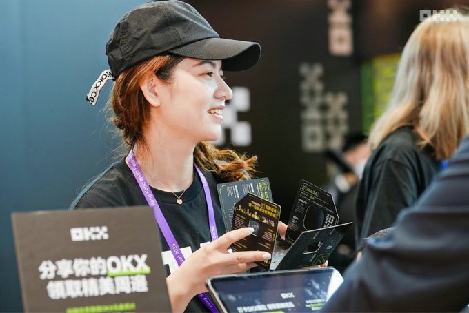 A smiling woman wearing a black cap holds a stack of OKX promotional cards at what appears to be a crypto event booth, with signage reading "分享你的OKX 领取精美周边" (Share your OKX, receive exclusive merchandise). The image captures the on-the-ground product marketing reality described in the post—engaging with potential users face-to-face, as OKX builds its brand presence and foundational infrastructure within the crypto community.