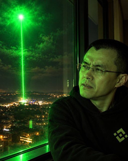 A man stands before a window overlooking a city at night, illuminated by an ominous green laser beam shooting skyward, with Binance's logo visible on his jacket—aligning with the post "It's coming," suggesting anticipation of a major Binance-related announcement or event.