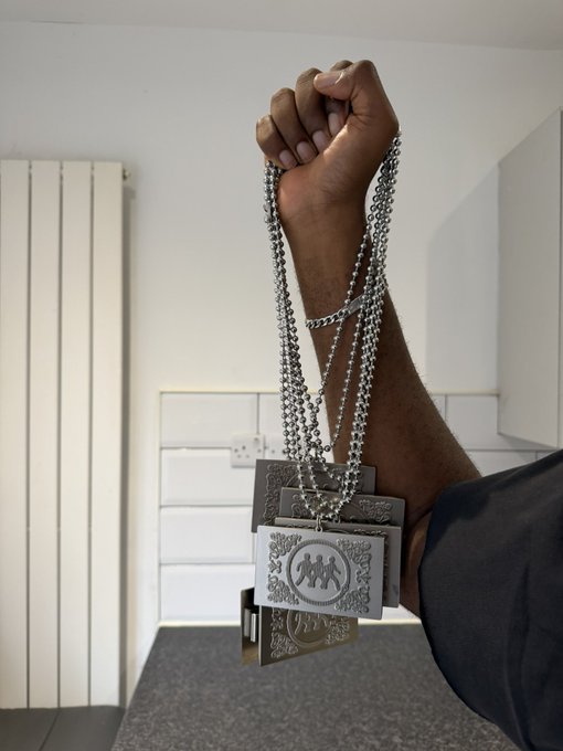 A hand proudly displays a custom wallet chain featuring multiple silver-toned metal pendants, including one engraved with a stylized "M" monogram and ornate decorative plaques, hanging from chunky silver links. The intricate jewelry piece showcases the finished product of the handcrafted wallet chain, emphasizing its bold, statement-making design and personalized detailing.