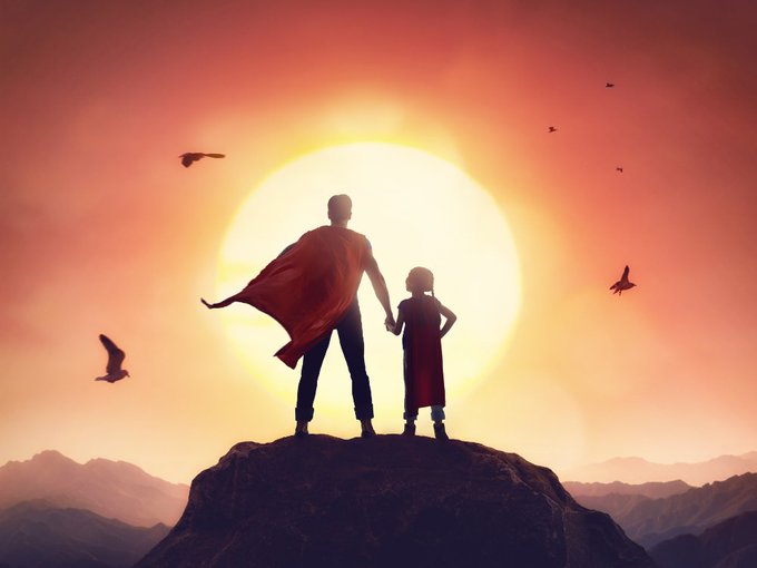 A silhouette of a man in a cape holding hands with a child stands atop a rocky hill, gazing out at a massive, bright yellow sun with birds flying in the orange sky.