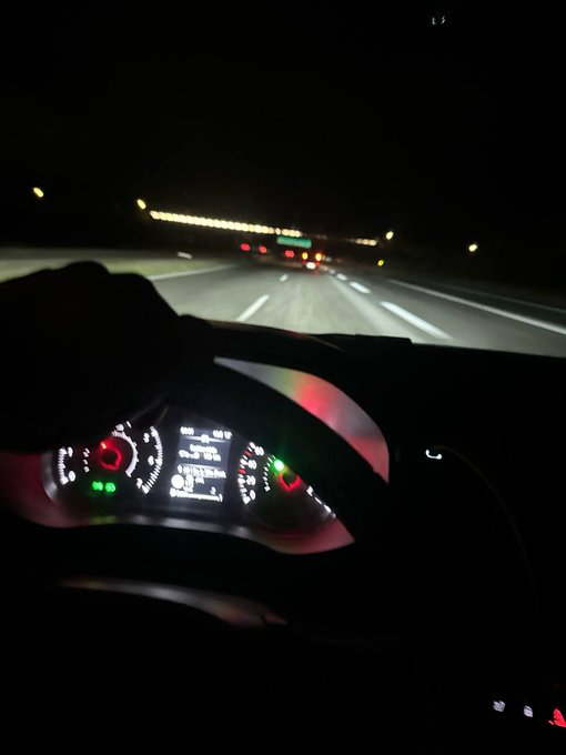 A nighttime photo taken from the driver's perspective inside a car, showing the illuminated dashboard with glowing green and red gauges, with the road ahead lit by headlights and overhead tunnel lights in the distance.