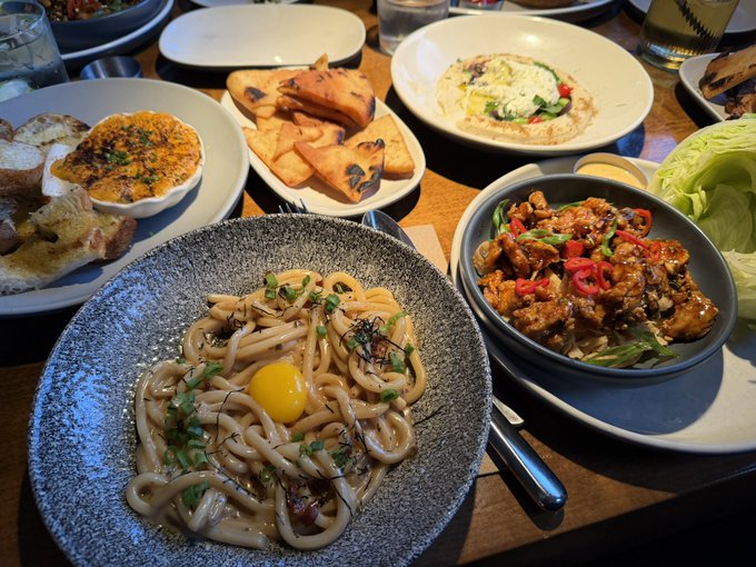 A spread of Asian-inspired dishes fills the table, including udon noodles topped with a raw egg yolk, crispy fried chicken garnished with chili peppers, flatbread, and a small gratin, perfect for a flavorful weekend meal.