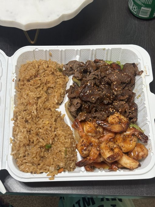 A takeout container holds a meal of fried rice, sesame beef, and shrimp, placed on a dark table next to a green can and a marble-patterned dish.