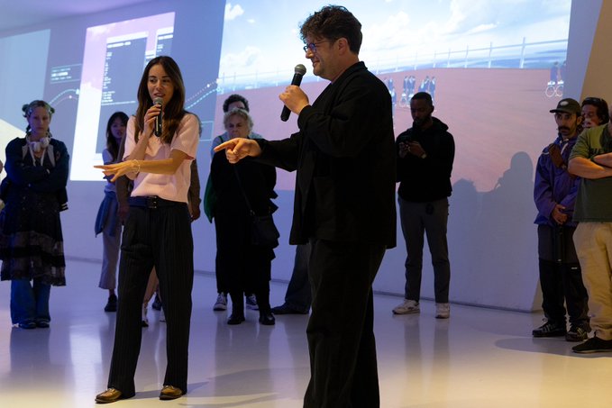 A woman in a pink blouse and black pants and a man in a black jacket both hold microphones and gesture while presenting in front of an audience at an indoor event.