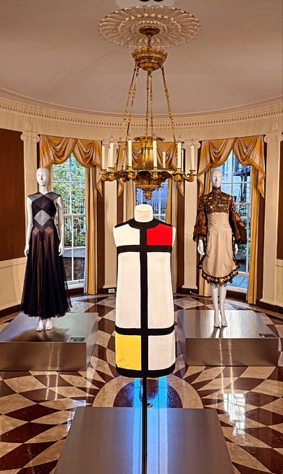 Three mannequins display vintage dresses inside an elegant room featuring an ornate chandelier, decorative crown molding, draped curtains, and a patterned marble floor.